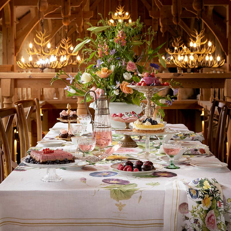 An elegant dining table set features a floral cloth, Koustrupcos Napkins: A Flower Garden by Jim Lyngvild, an array of desserts, and a large bouquet. Warm lighting and a wooden interior enhance the cozy ambiance, echoing Jim Lyngvilds distinctive style.