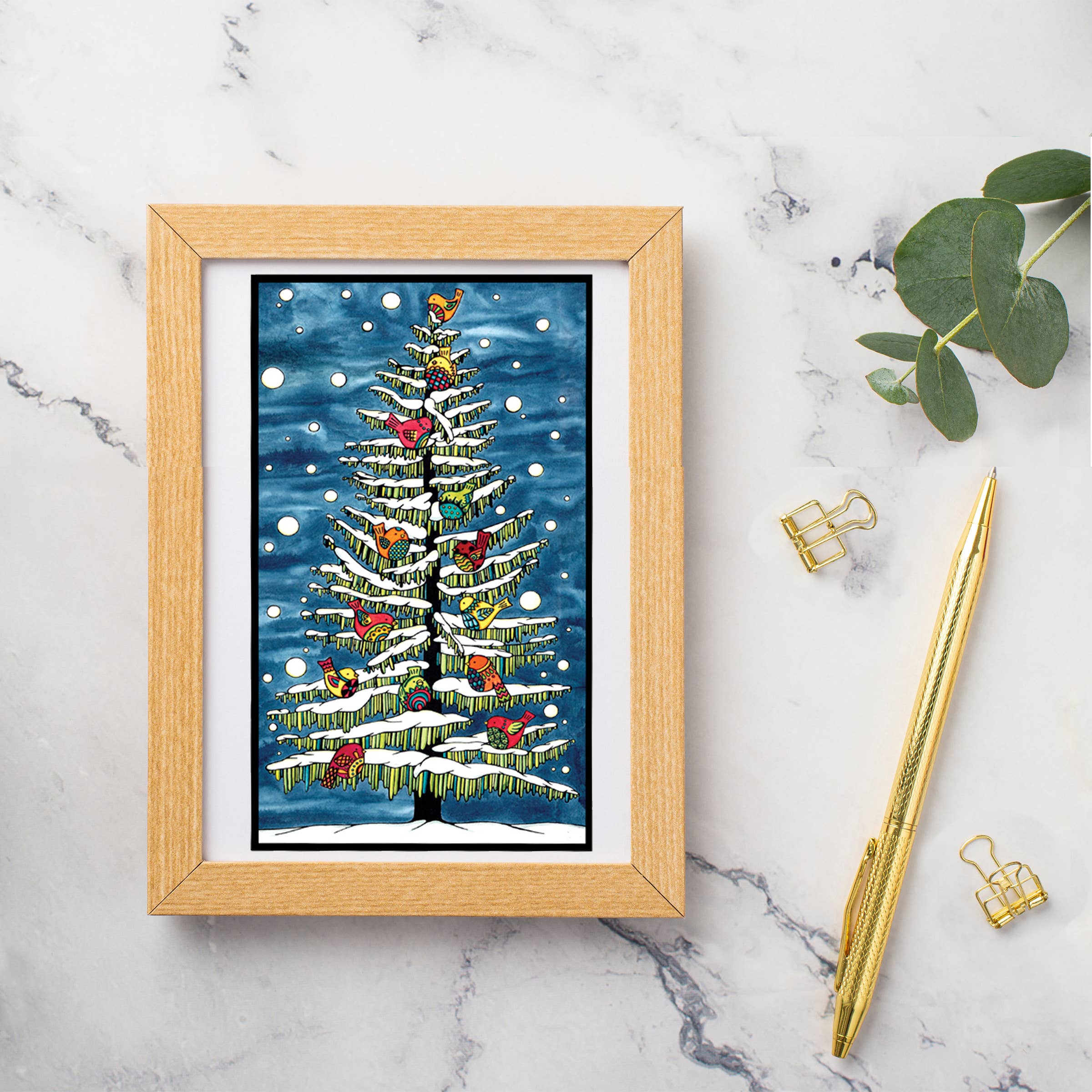 The "Holiday Birds in Snow" Greeting Card by Sarah Angst (5x7, blank inside, with envelope) features festive birds in a Christmas tree. Shown framed on marble with gold pen, binder clips, and leafy stem—perfect for sending holiday cheer.