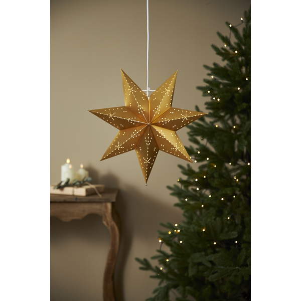 The Star Trading Classic Hanging Star Gold (16.5") glows near a Christmas tree with white lights. Candles and gifts on a wooden table add to the cozy holiday decor.