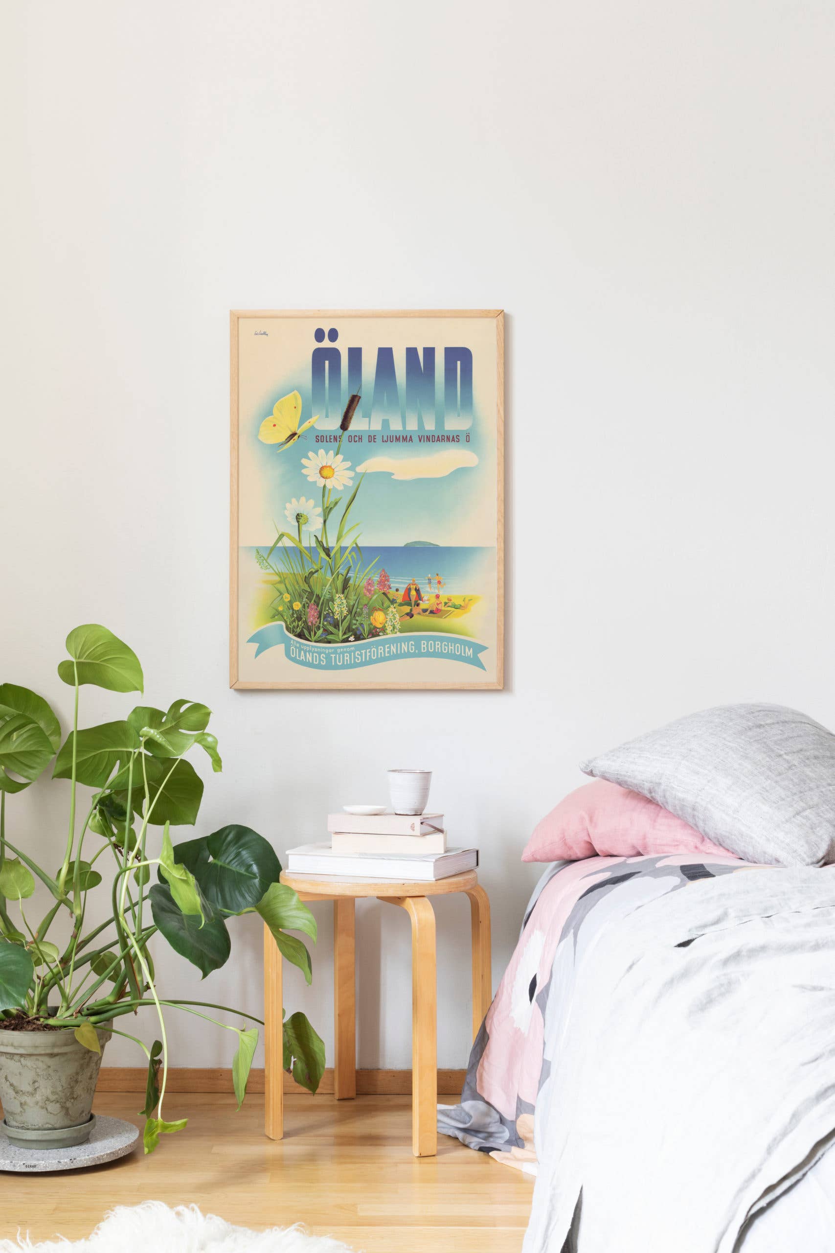 A bedroom corner includes a vintage Öland Scandinavian Travel Poster by Lindblom, a wooden side table with books, a glass, and a candle next to a bed with pink and gray sheets. A potted plant adds vitality to this curated space.