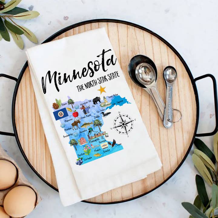 A delightful Tea Towel: Minnesota State Map Souvenir Kitchen Cotton Flour Sack Towel, showcasing a design labeled The North Star State, is gracefully arranged on a round wooden tray with measuring spoons and eggs nearby—an ideal housewarming gift.