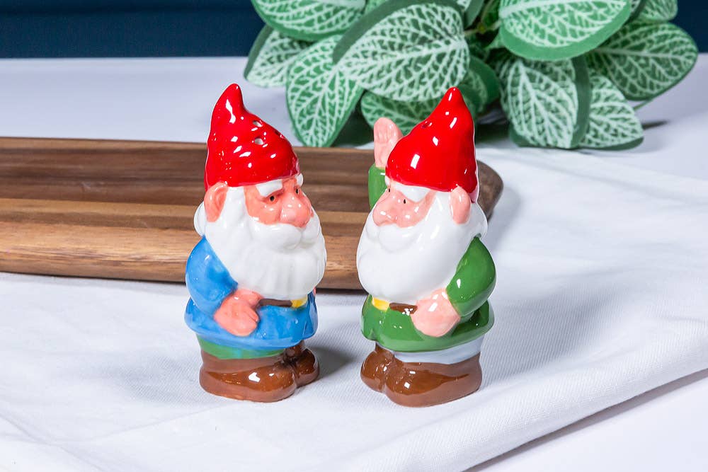 The Shakers: Garden Gnomes - Salt & Pepper Shakers feature two ceramic gnomes with red hats, one in a blue coat and one in green, displayed on a white cloth near a wooden tray and a leafy plant.