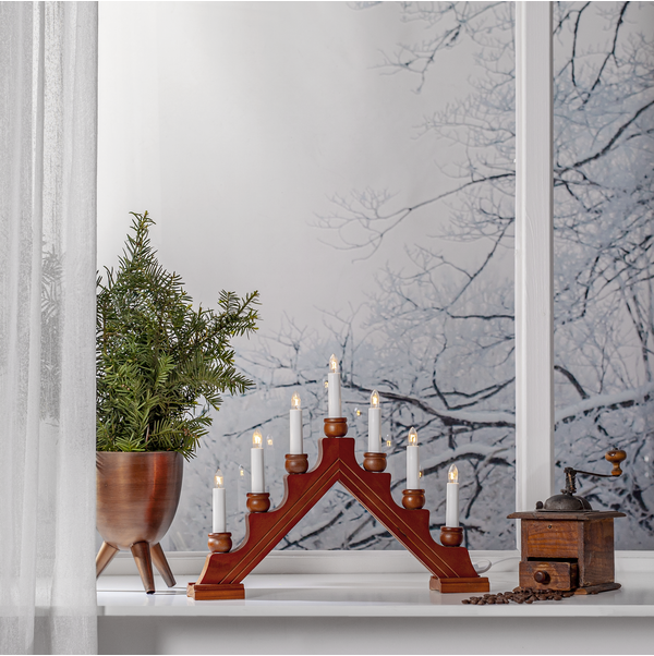 A Star Trading Karin Electric 7-Light Candelabra in red, brown, or gray glows on the windowsill next to a potted green plant and coffee grinder, framing the snowy trees outside.