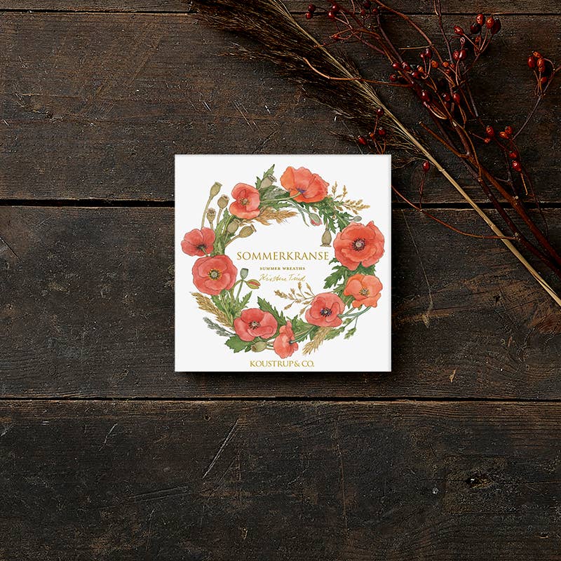 A square card folder sits on wood, featuring a floral wreath of red poppies and green leaves. Named Card Pack: Summer Wreaths by Koustrup & Co - Denmark, it is elegantly adorned with a branch of red berries for added charm.