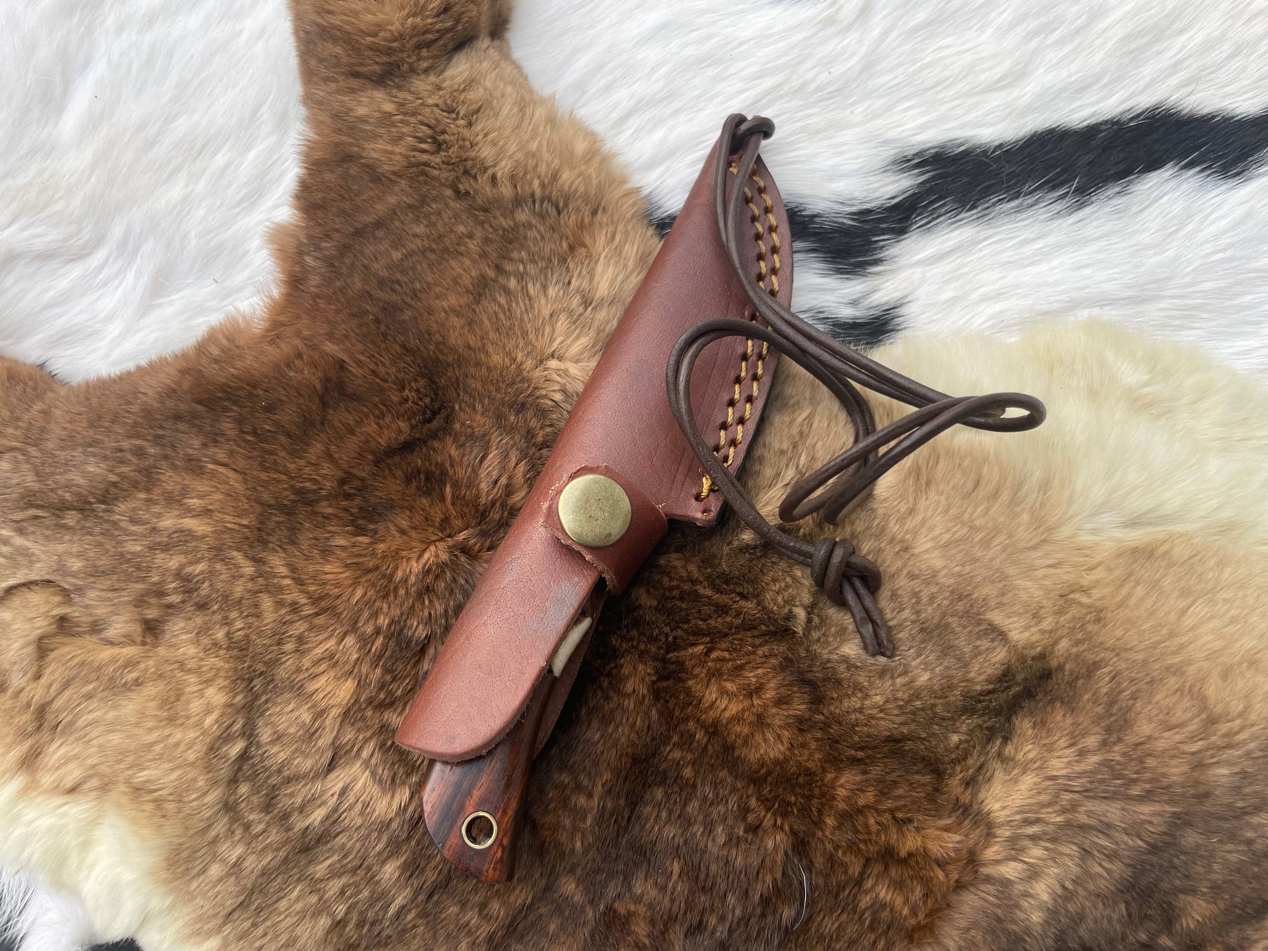 A brown leather sheath with a snap button and lanyard rests on a fur pelt background, perfect for securing your Damascus Steel Skinner Knife with Rosewood & Stag Grip.