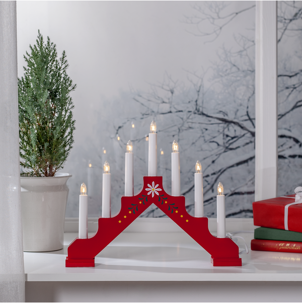 The Star Trading Adam 7 Bright Red Floral Candelabra with seven glowing candles sits on a white windowsill beside a potted evergreen and wrapped gifts, creating charming window decor against the snowy backdrop.