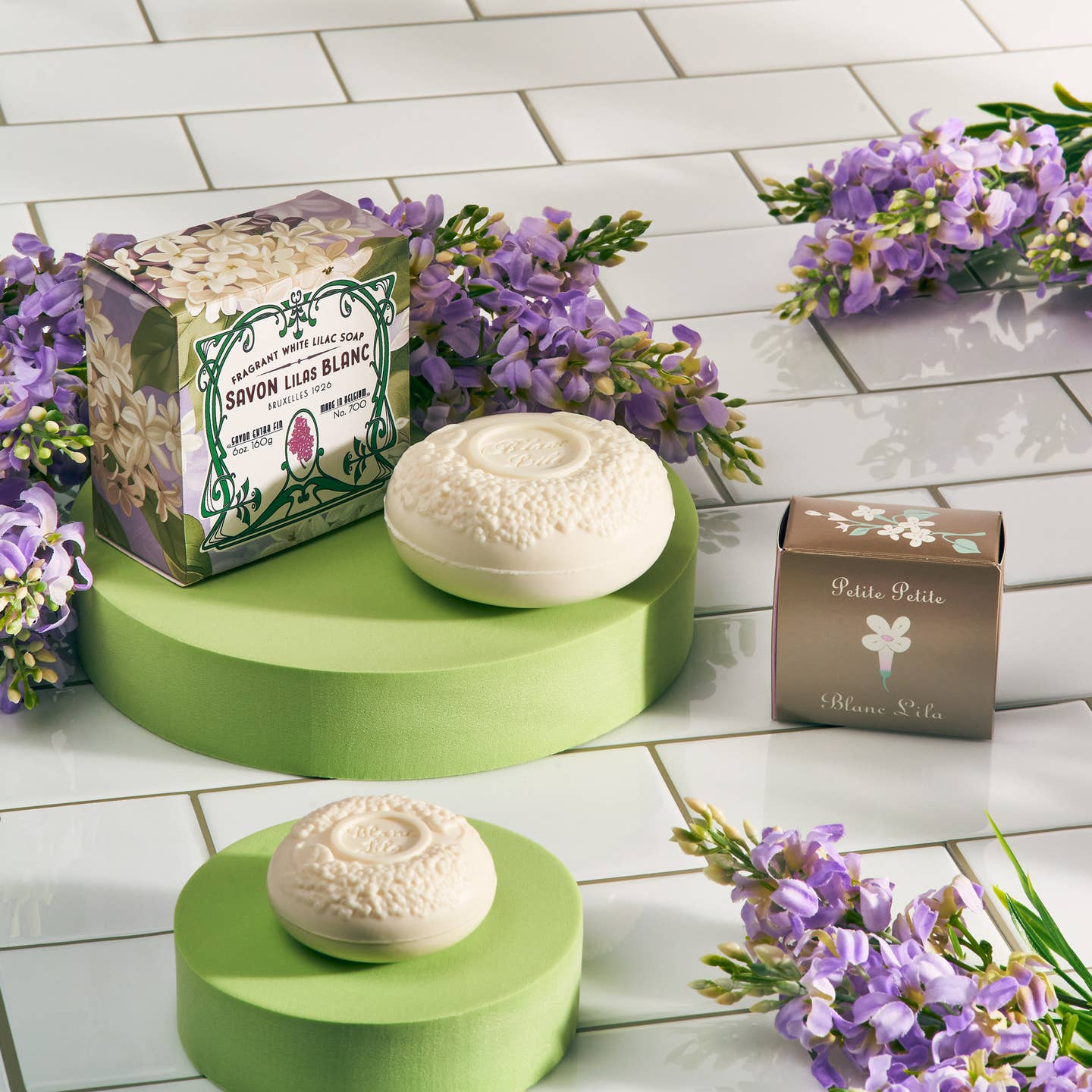 Two Blanc Lila Lilac Savon Bar Soaps with embossed designs rest on green platforms with floral packaging and purple flowers atop white tiles. This vegan soap brings a touch of elegance to any space.