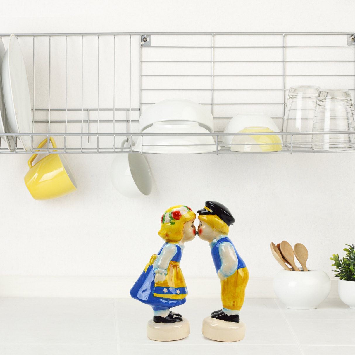 The Swedish Couple Salt & Pepper Shakers with Magnets stand on a kitchen counter, accompanied by a dish rack above, which holds plates, cups, and glasses.