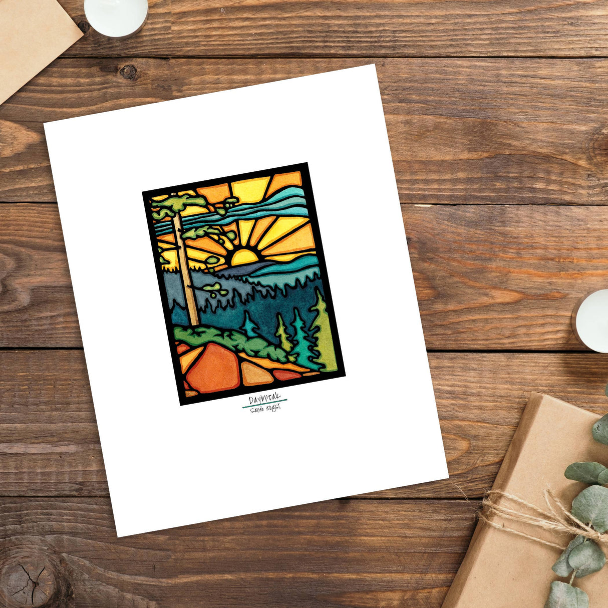 The Artwork: Daybreak Print 11"x14" by Sarah Angst features a stained glass-style sunrise over mountains, trees, and a path. Set with candles, an envelope, and a package, this print adds warmth to any space.