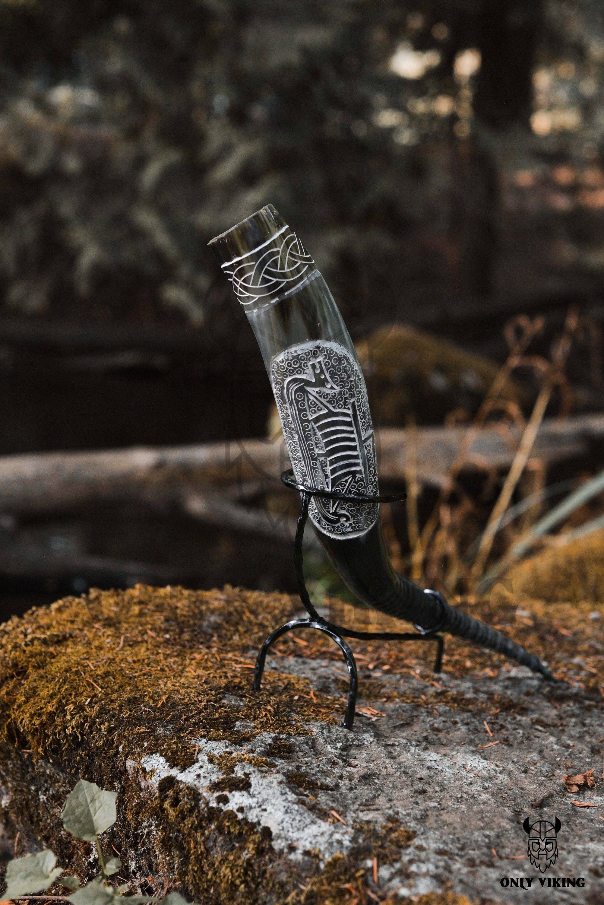 The Ravens Viking Ship Handcrafted Drinking Horn (14-18 inches) rests on a metal stand outdoors atop a mossy rock.