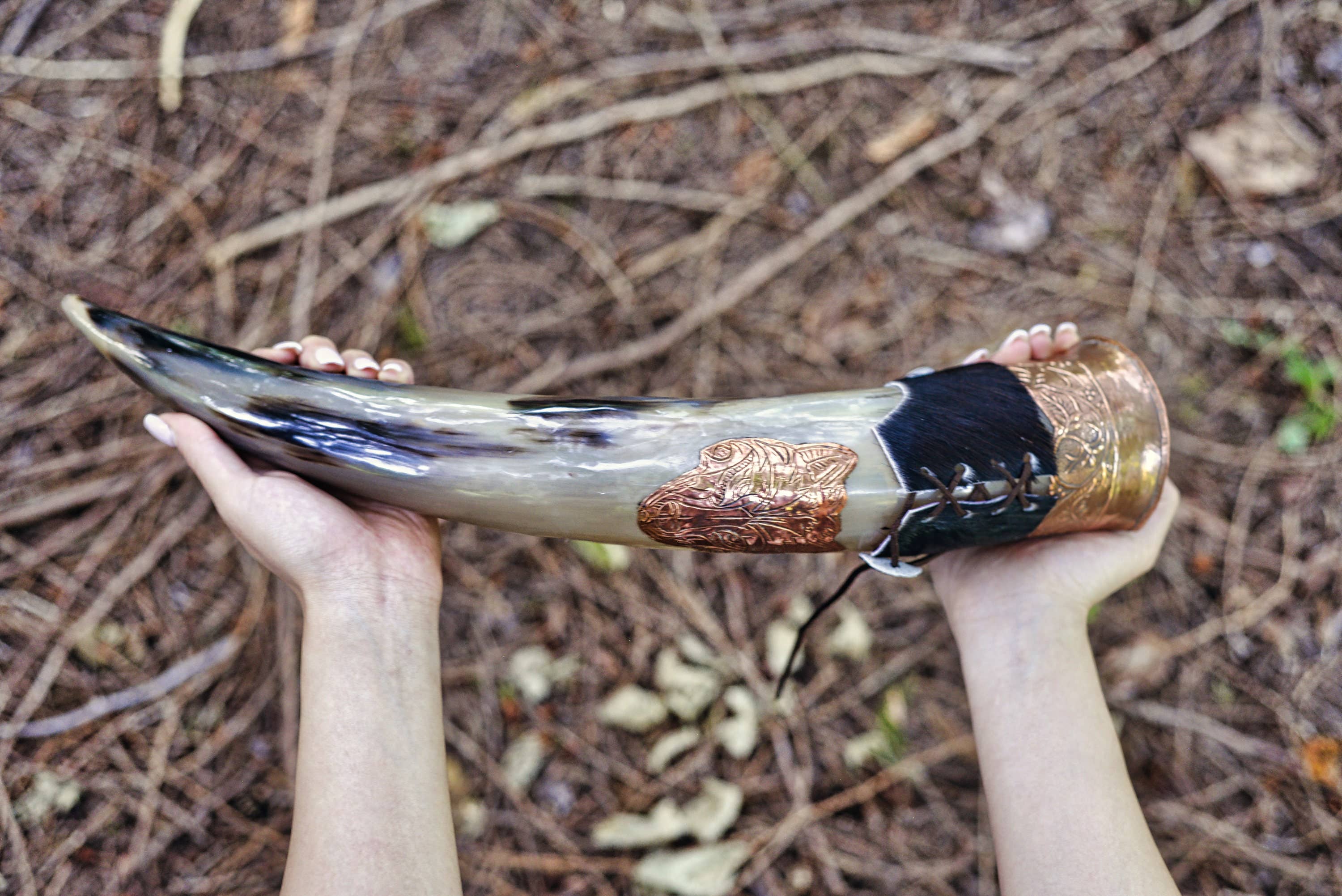 A person holds the Drinking Horn: Authentic Viking Wedding Gift, featuring metal accents and carved designs, above a forest floor covered with leaves and twigs.