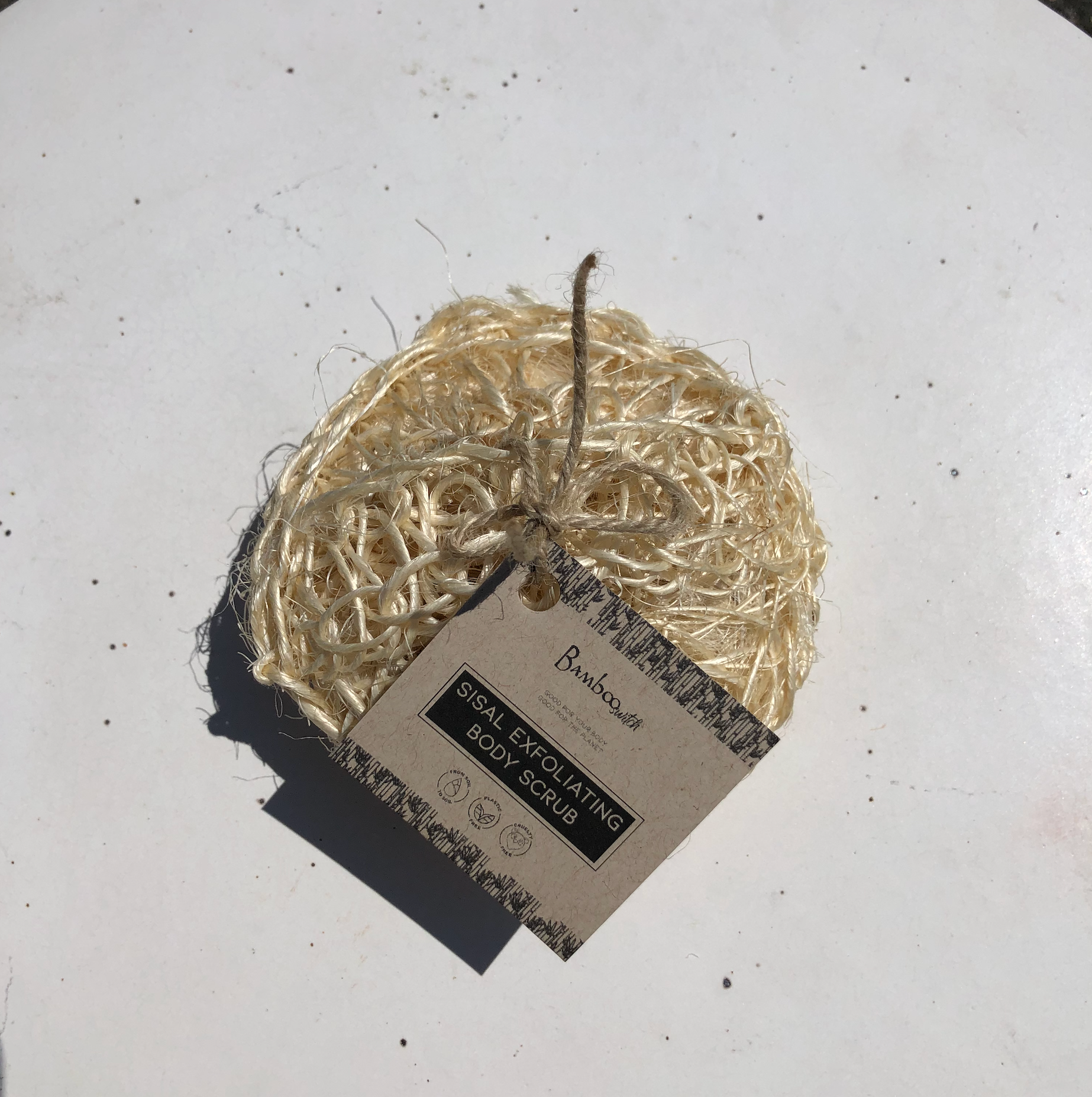 The Scrub: Sisal Exfoliating Body Scrub is a round, eco-friendly exfoliator with a label and twine, shown on a white surface in sunlight—ideal for zero-waste beauty routines.