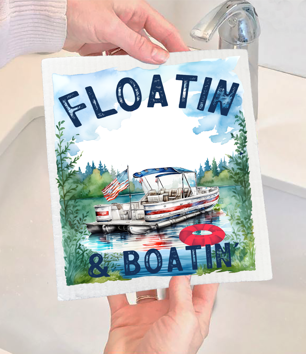 A person holds the Dish Cloth: Floating and Boating on the Lake Swedish Dishcloth, which features a pontoon boat on a lake, an American flag, and bold blue text reading FLOATIN & BOATIN.