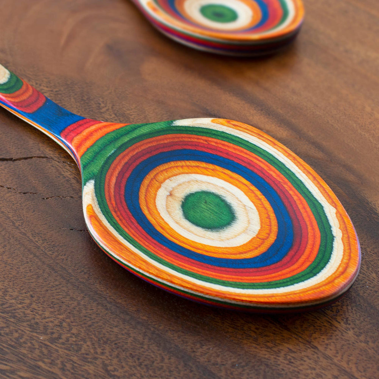 Birch serving spoon with green, orange, blue, red, and yellow rings on handle lying on a brown wooden surface; another birch spoon is partially visible in the background.
