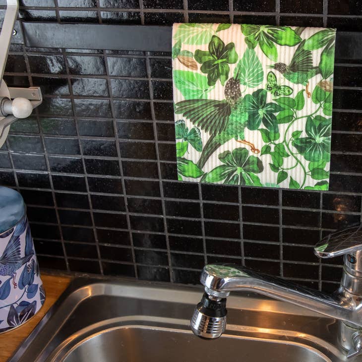 A Dish Cloth: Green Birds in Trees hangs on a black tiled backsplash above a stainless steel sink and faucet, showcasing its eco-friendly, biodegradable charm.