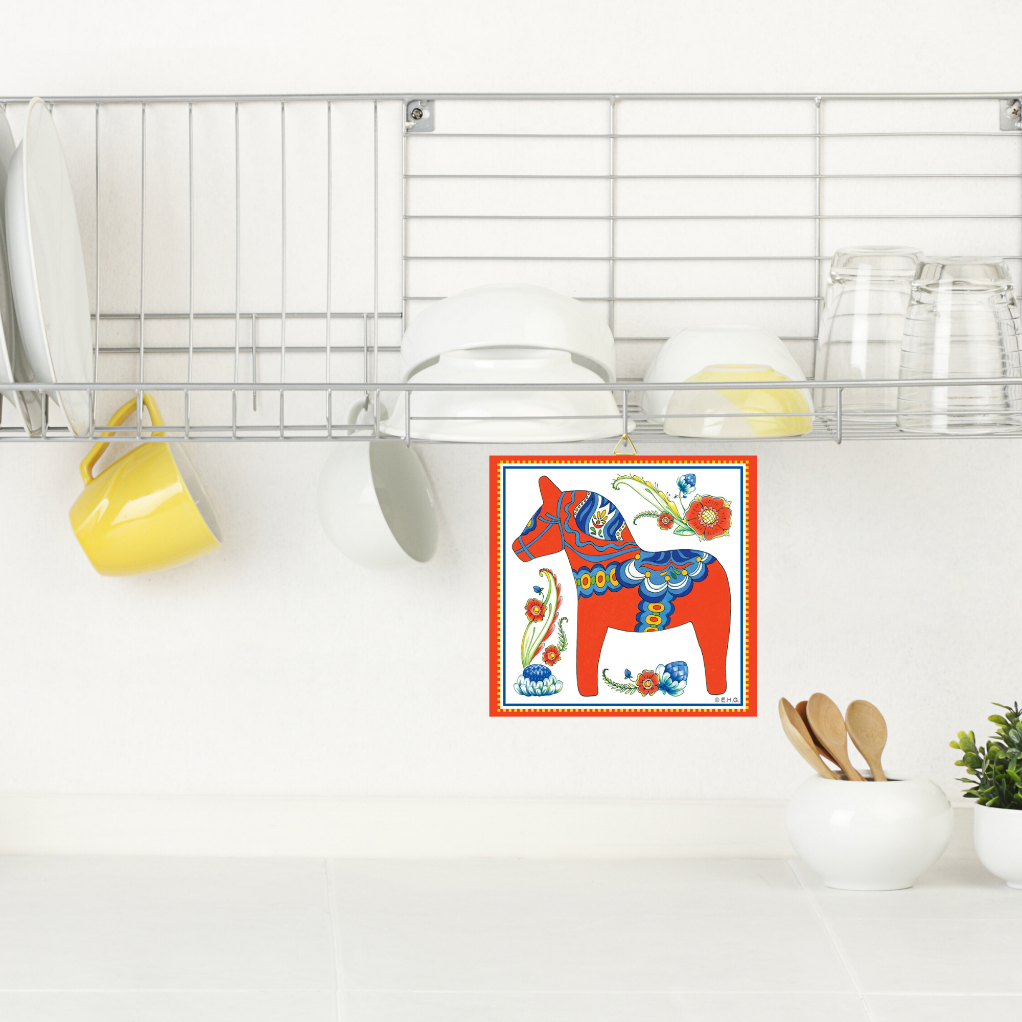 A kitchen shelf holds white bowls, plates, glasses, and yellow mugs above a counter with utensils. On the wall hangs a Red Swedish Dala Horse Wall Decor Tile (6x6), adding vibrant color to the space.