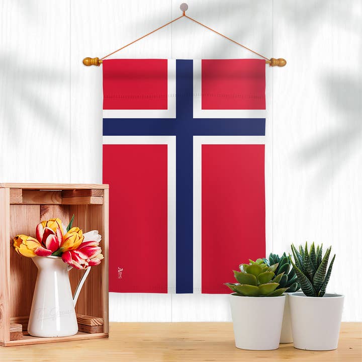 The Flag: Norway Garden Flag - Norwegian Nationality Home Decor Flag hangs on a white wall above a table with a wooden crate, vase of tulips, and two small potted succulents, celebrating Norwegian heritage.