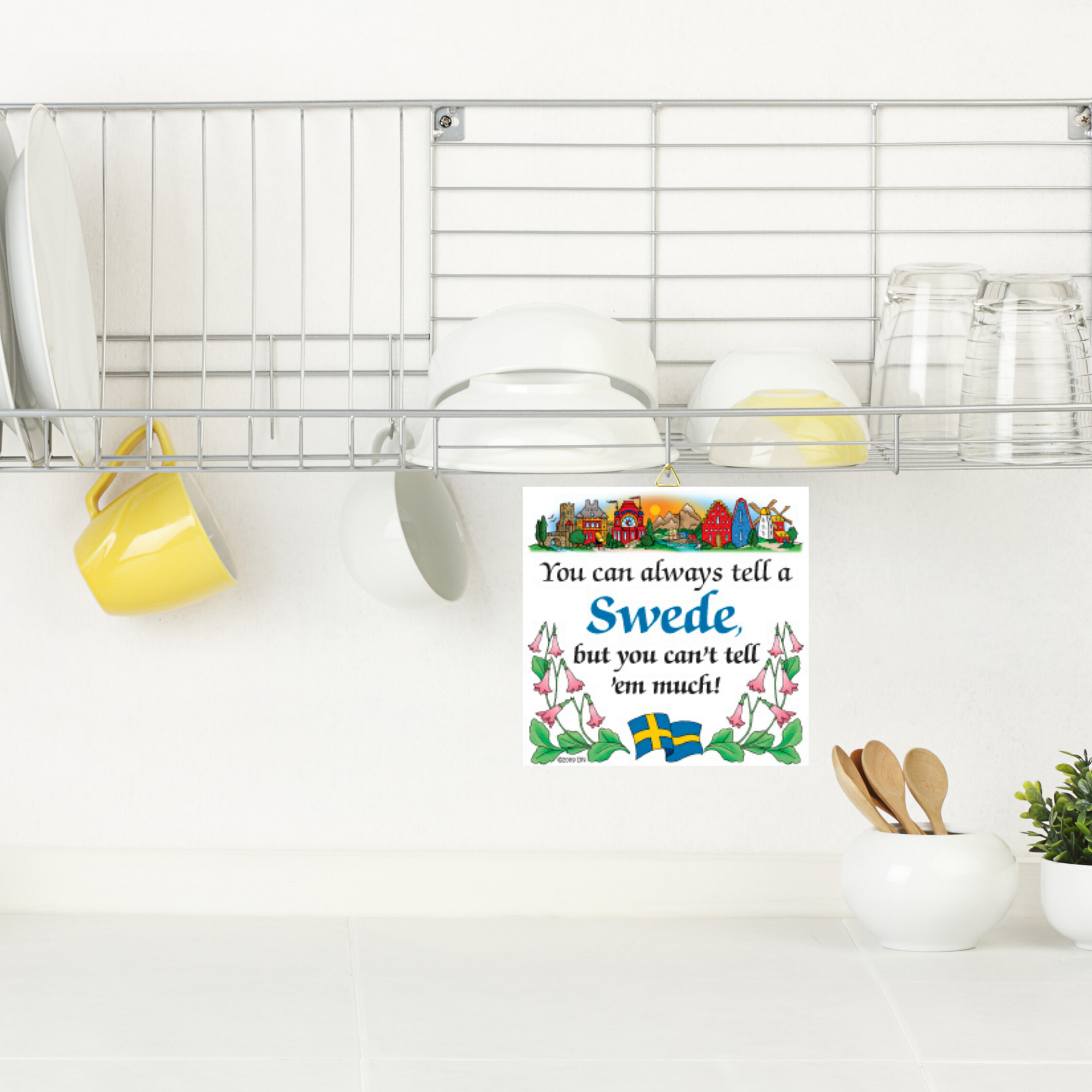 Above the counter, a dish rack holds plates and mugs. Below, the 6 Tile: You Can Always Tell A Swede, But You Cant Tell em Much! with hanger features floral, Swedish flag, and Dala horse accents for wall decor.