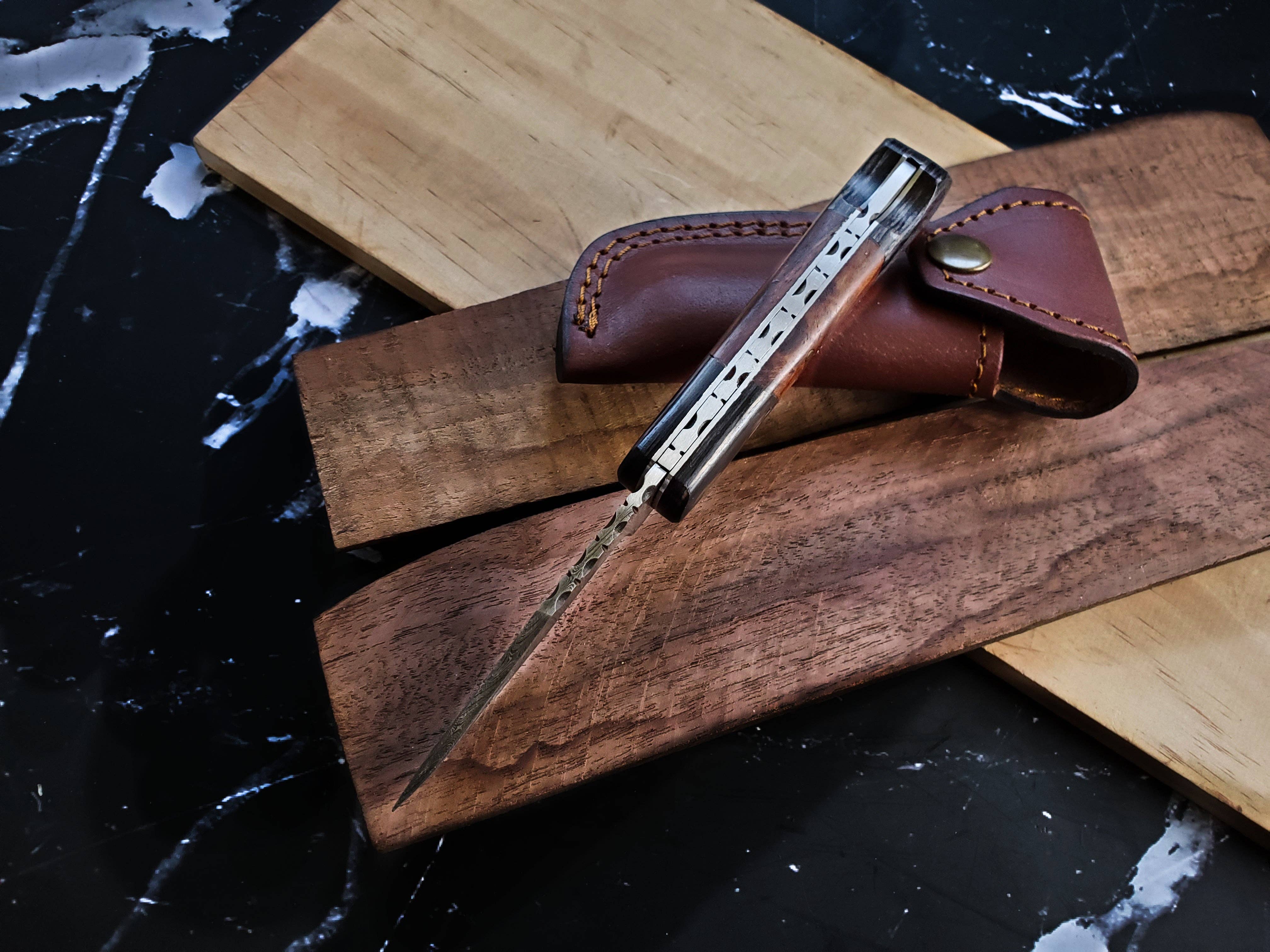 The Knife: Damascus Steel Pocket knife with a decorated spine and walnut handle sits on wooden boards beside its brown leather sheath atop black marble.