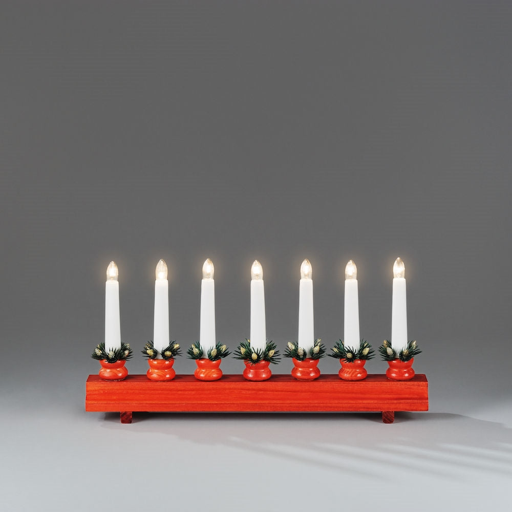 The Konstsmide Candelabra - Ingrid features seven white electric candles, each nestled in a red stained base with green accents, set against a gray background.