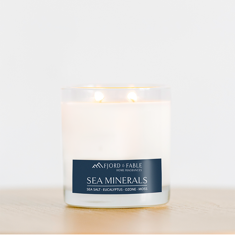 A lit Fjord & Fable Sea Minerals 4oz candle with 2 wicks and a blue label rests in a clear glass bowl on a light wooden surface; it offers 20+ hours of burn time.