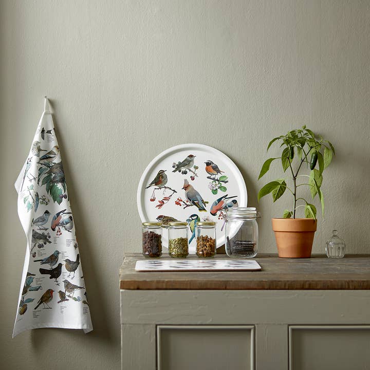 A wooden counter holds three glass jars, a potted plant, and a dish with bird illustrations. Next to it, a Garden Birds tea towel by Carl Christian Tofte (100% organic cotton, 19x27, Koustrupco) hangs on a hook against a plain wall.