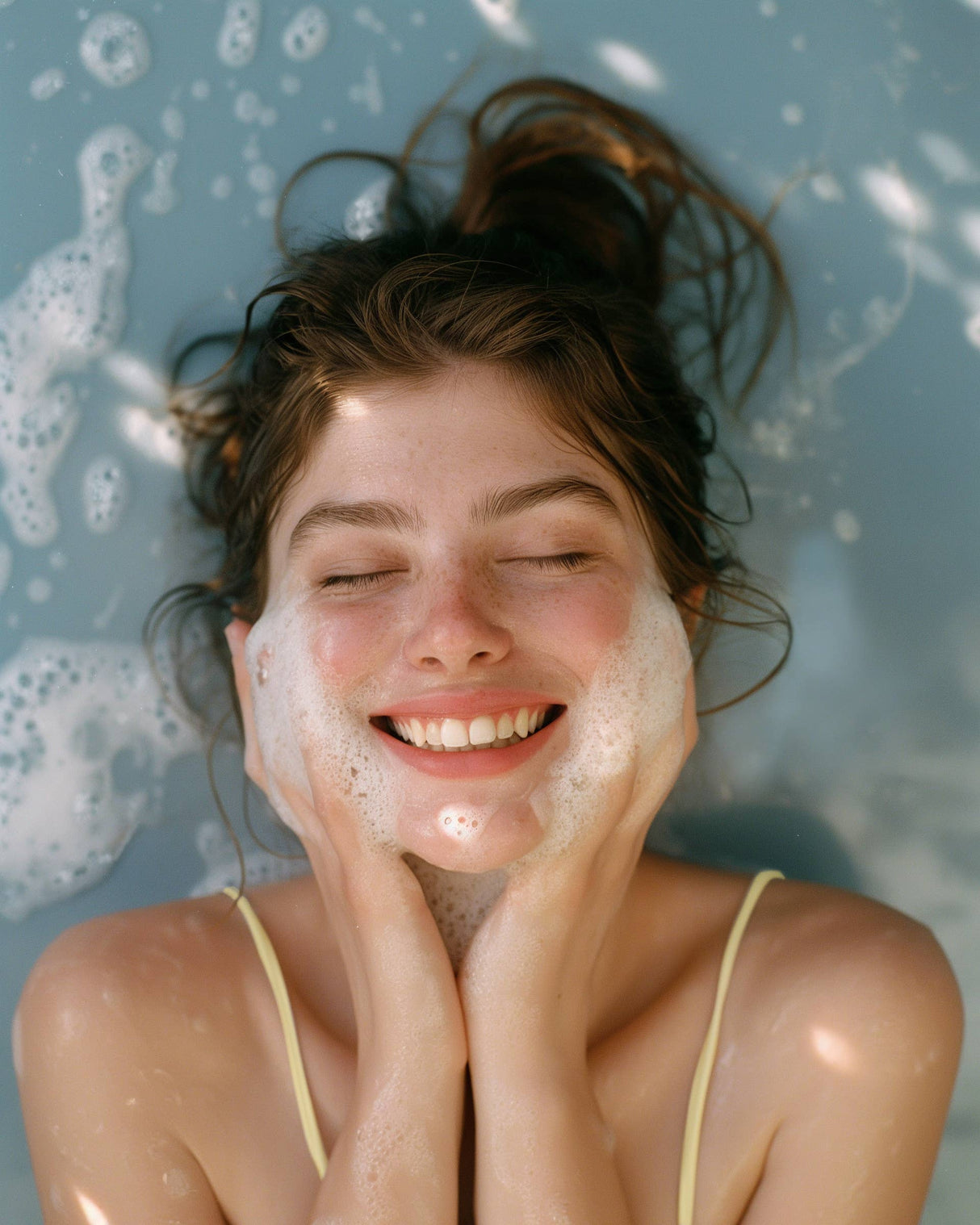 A young woman with closed eyes smiles while washing her face, holding the Eggwhite & Chamomile Facial Soap from the Gingham Gift Box (1.76 oz), lying back in water with bubbles.