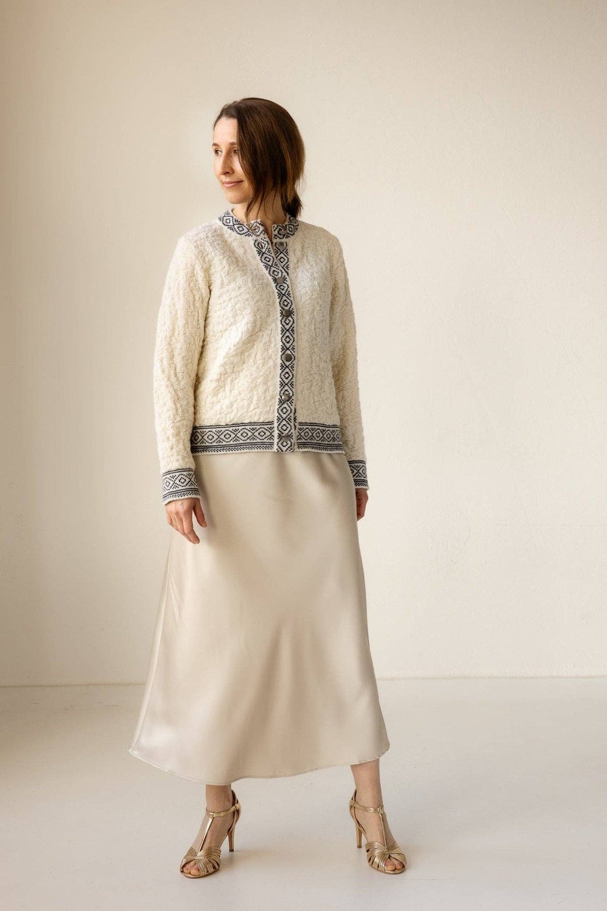 A woman is indoors wearing the Bryggen Women's Norwegian Cardigan (White), a long beige satin skirt, and gold open-toe heels.