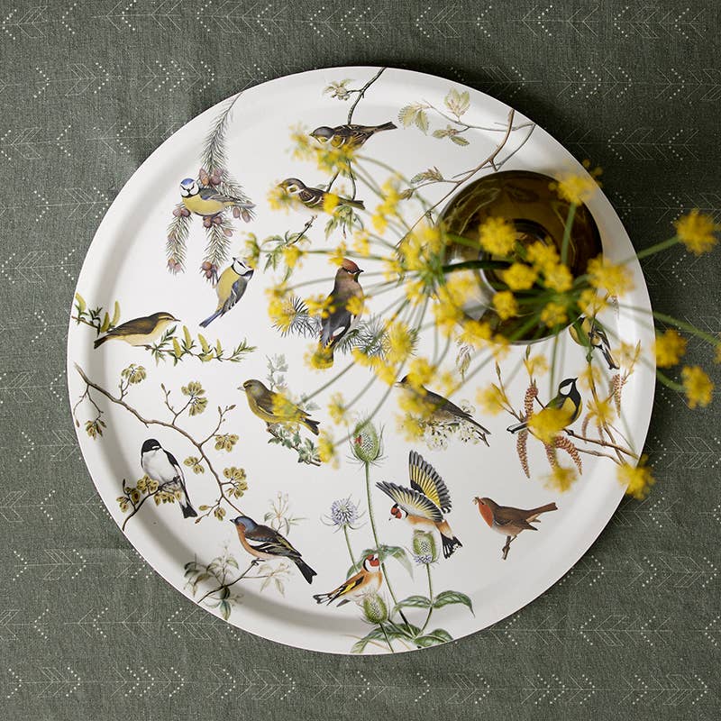 The 14.9 Garden Birds birch veneer serving tray by Koustrupco of Denmark, featuring colorful bird illustrations, sits on a green patterned cloth holding a glass vase with yellow flowers.