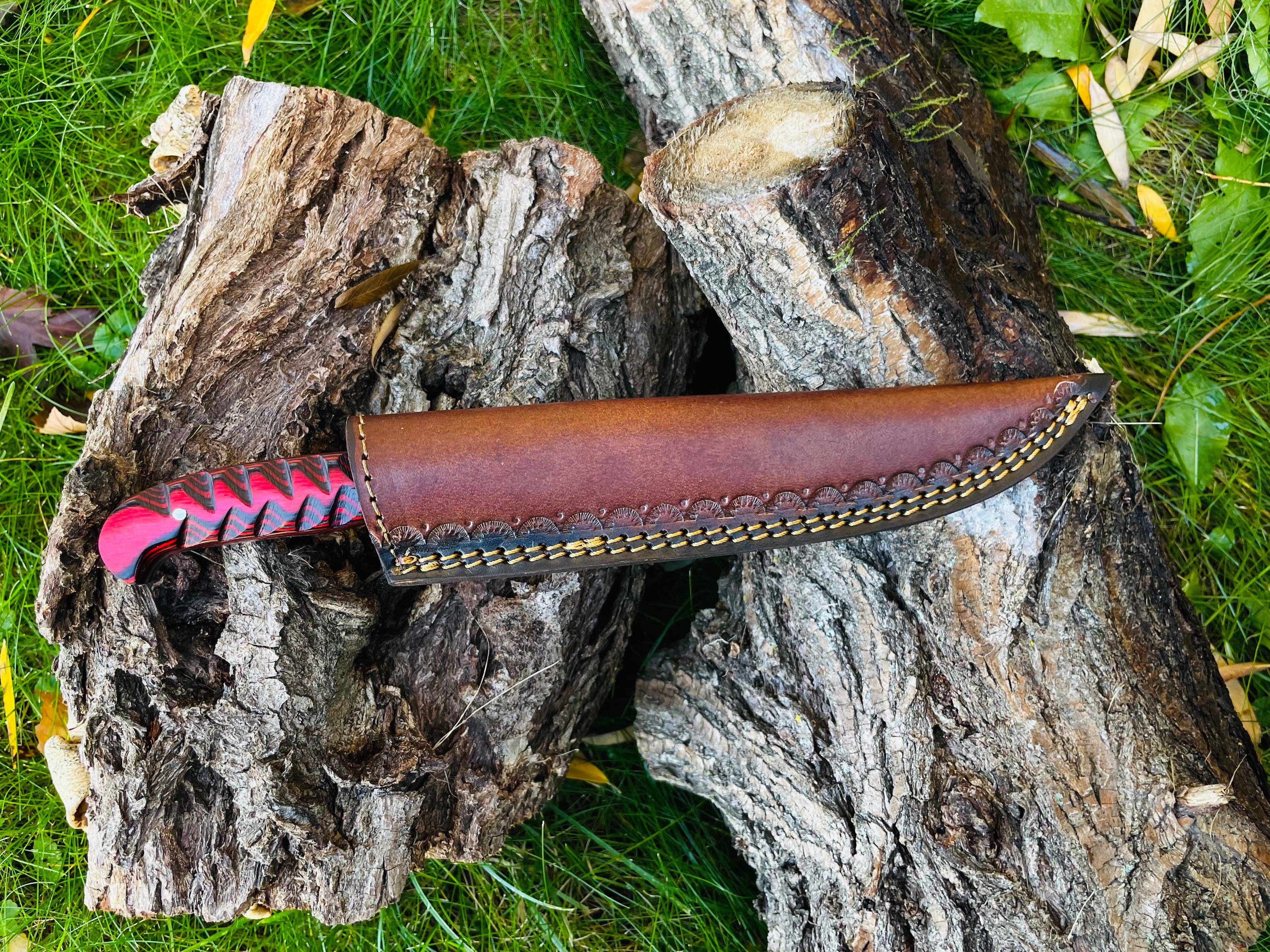 The Knife: Damascus Steel Custom Flay Knife with carved red and black grips in a brown leather sheath rests on tree bark atop green grass.