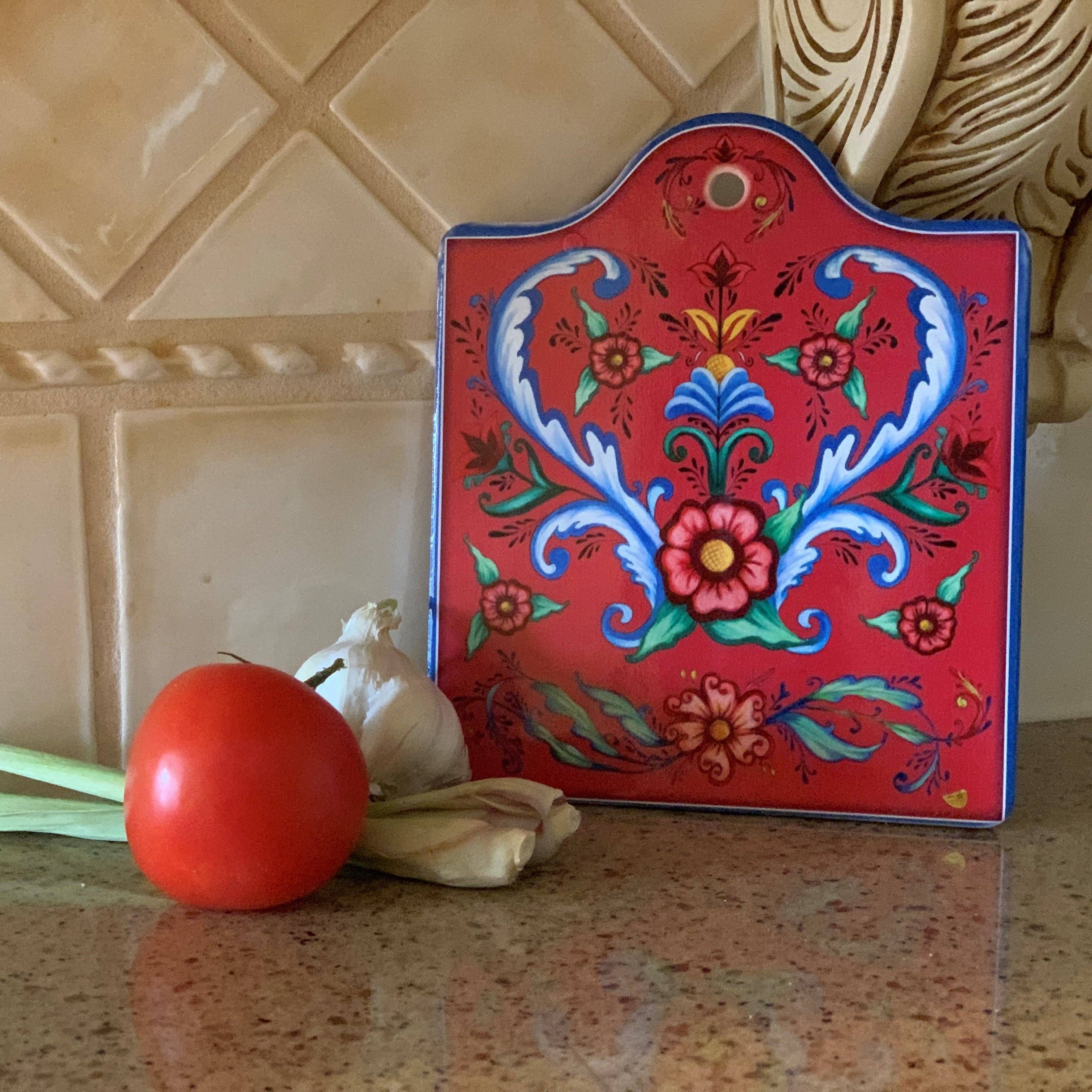 The Red Rosemaling Ceramic Cheeseboard with cork backing, featuring a Scandinavian floral design, stands on a kitchen counter beside a tomato, garlic bulb, and green onions.