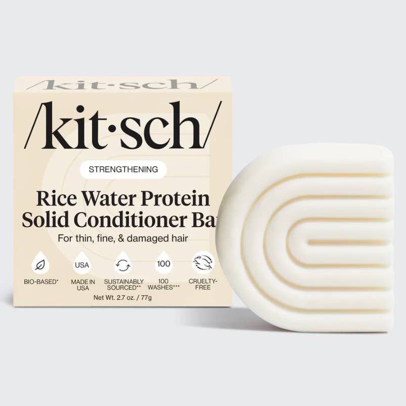 A box labeled "KITSCH Conditioner: Rice Water Protein Solid Conditioner Bar for Hair Growth 2.7oz" sits next to a white, curved, textured conditioner bar. The packaging highlights benefits for hair growth and added volume in thin, fine, and damaged hair—powered by Rice Tein NPNF—and displays icons indicating the product is bio-based, cruelty-free, and made in the USA.