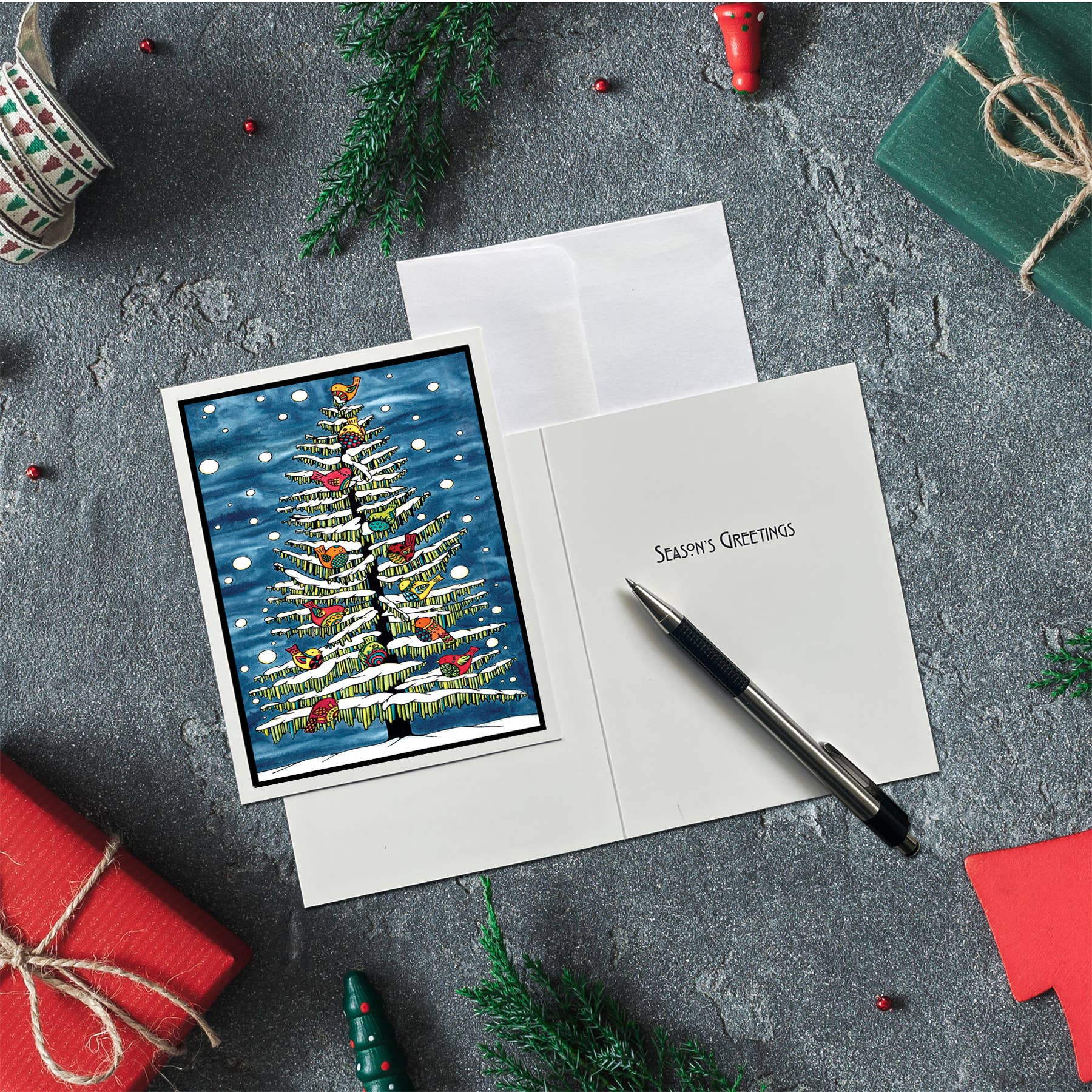 A packaged Card Pack: Birds in Snow on Tree Christmas card is open on a gray surface, with a pen inside and nearby wrapped gifts, ribbon, and greenery. Includes 8 cards and envelopes.