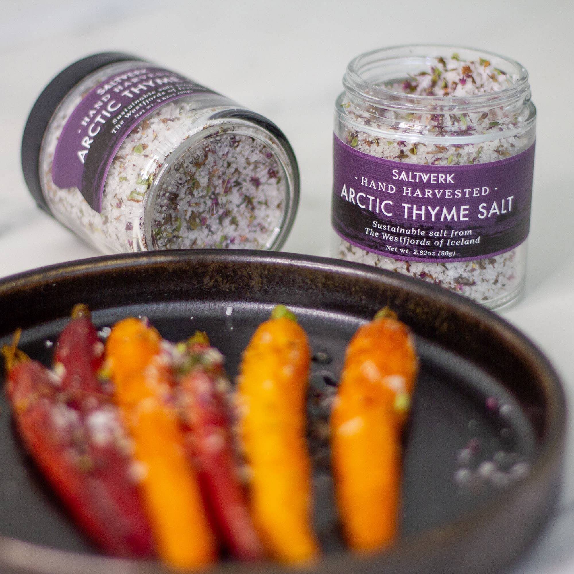 An open jar from the SALTVERK Gift Set (Flaky Sea Salt, Lava Salt, Birch Smoked Salt, Arctic Thyme Salt) lies beside an upright jar, while vibrant roasted carrots sprinkled with salt are arranged on a black plate in front.