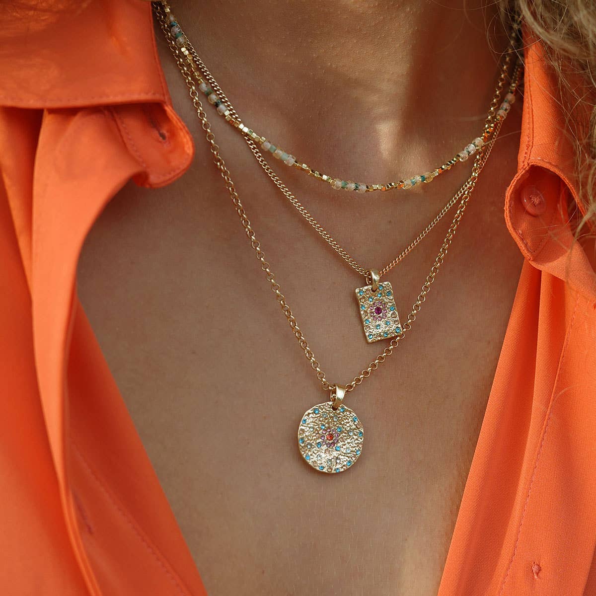 A person wearing an orange shirt and three layered gold necklaces, including a layering pendant and the A&C Oslo Vibrance Gold Coin Necklace with colorful stones.