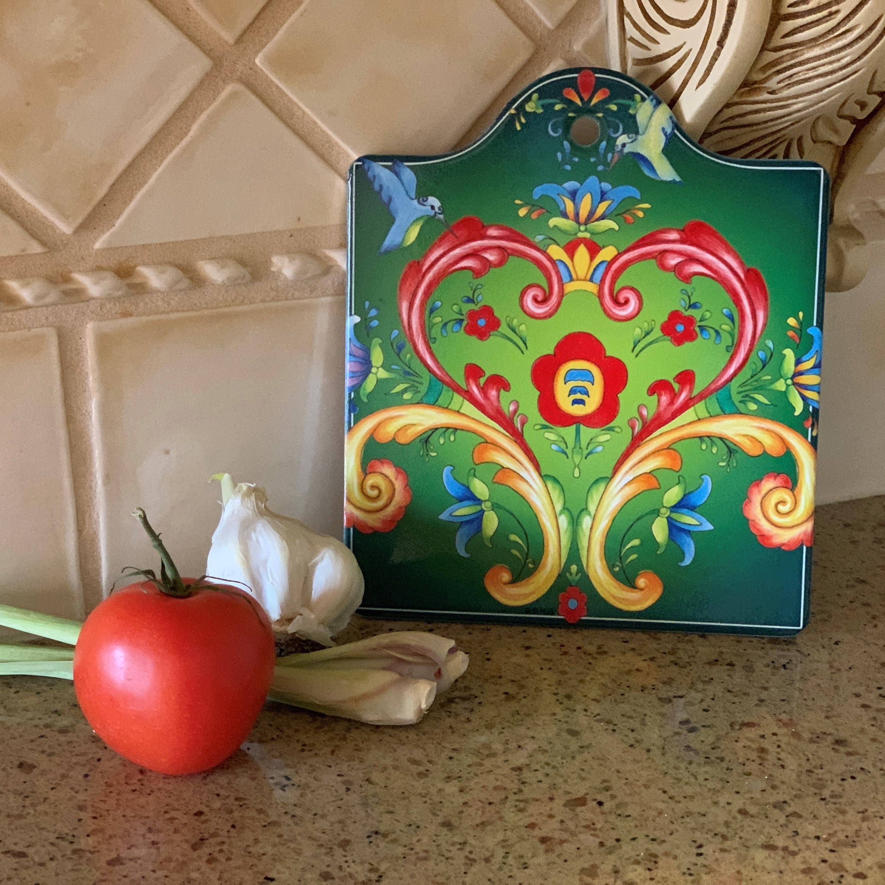 A tomato, two garlic cloves, and a stalk of lemongrass sit on a countertop next to a Rosemaling Green Ceramic Cheeseboard Kitchen Trivet, adding Scandinavian charm to the kitchen scene.
