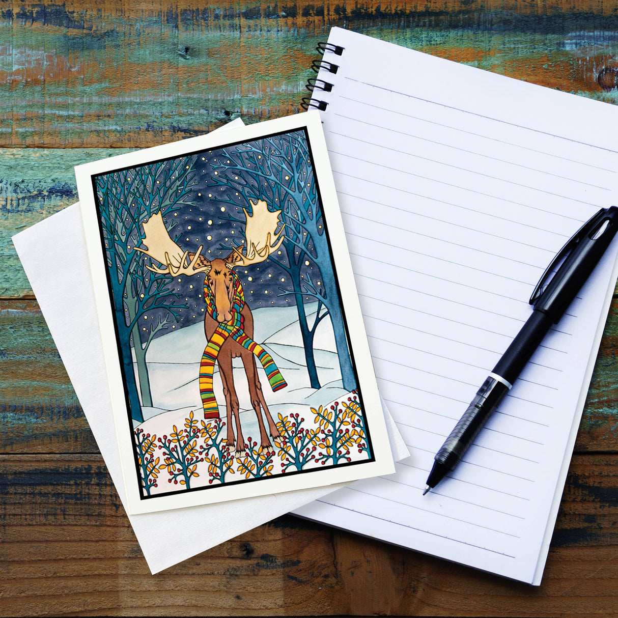 A Holiday Moose 5x7 greeting card by Sarah Angst, featuring a moose in a colorful scarf in a snowy forest, sits atop a blank notepad with a black pen on a wooden table. Card comes with envelope and is blank inside.