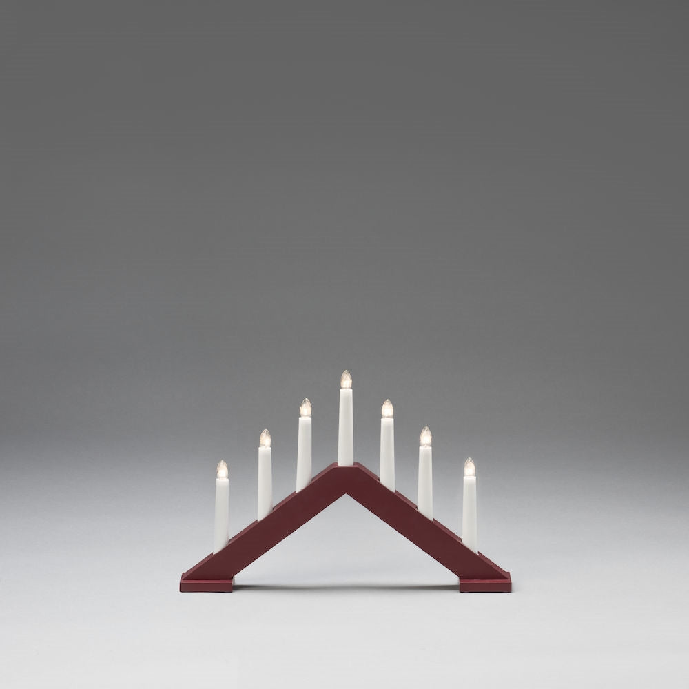 The Johanna electric candelabra by Konstsmide, in a dark red finish, features seven illuminated white candles arranged in a triangular configuration against a gray background.