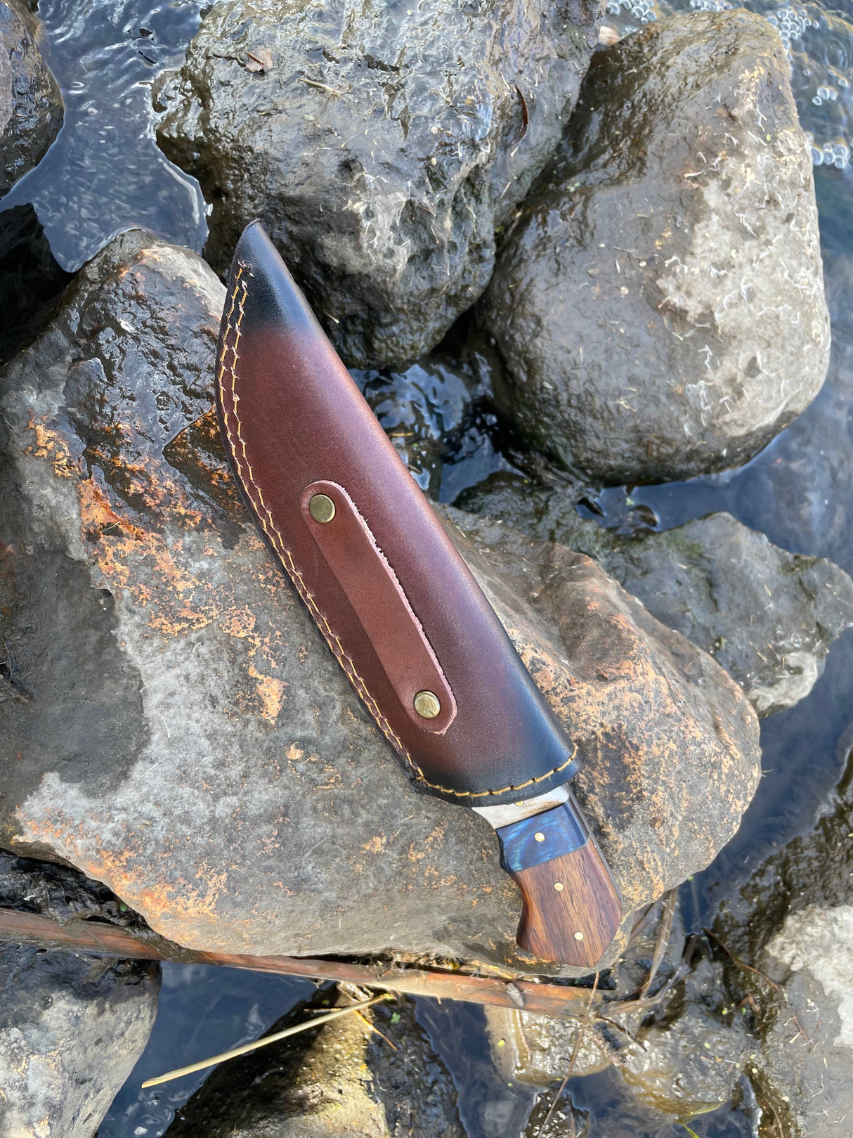 The Titan International Handmade High Carbon Damascus Steel Hunting Knife, featuring a Diamond Wood and Stag Horn handle plus leather sheath, rests on wet rocks by a stream—an essential for any outdoor adventure.