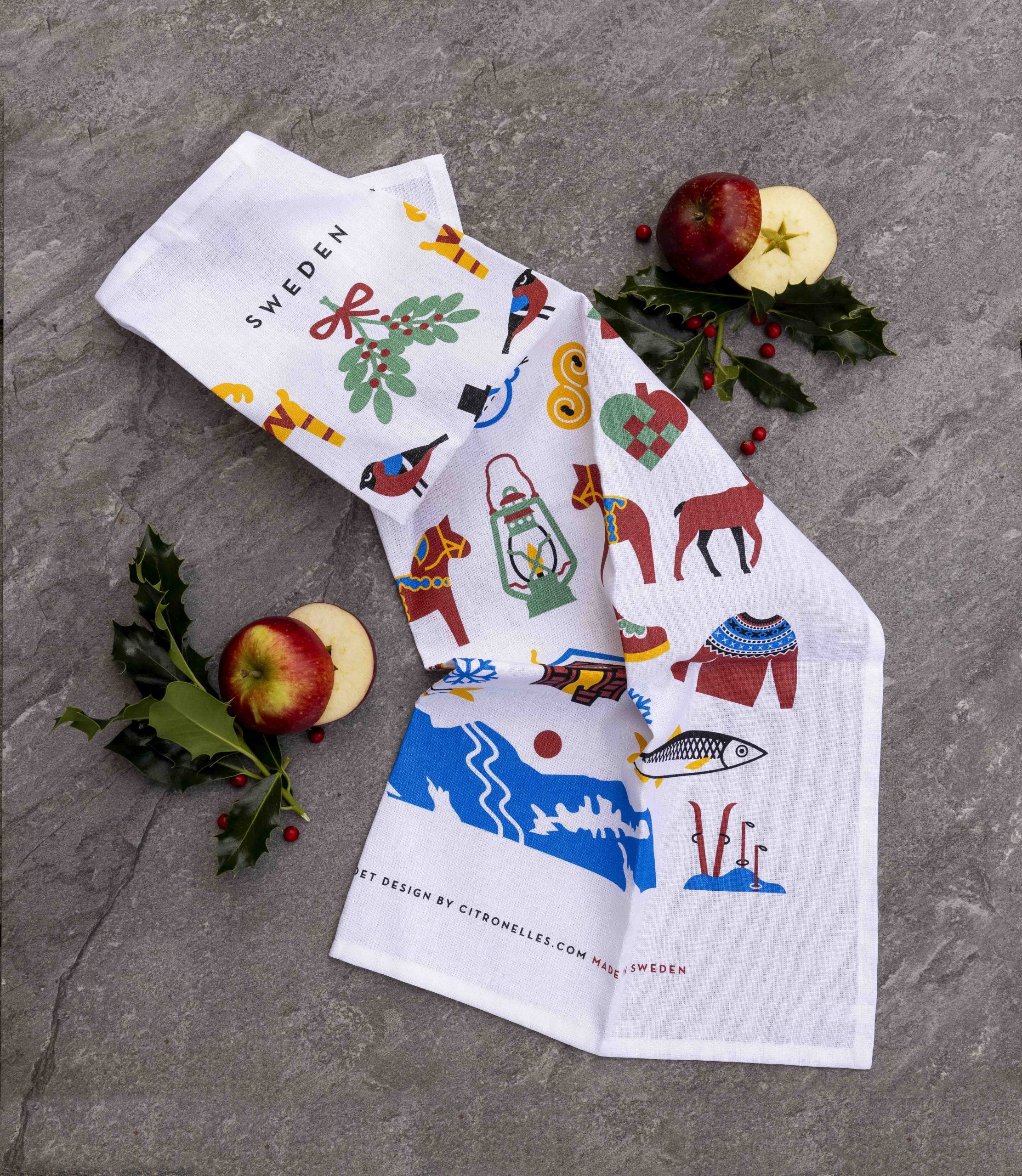 The Swedish Winter Traditions Tea Towel (50 × 70 cm) in a white cotton/linen blend features colorful Scandinavian designs—ideal for kitchen decor and displayed on gray stone with apples and holly.