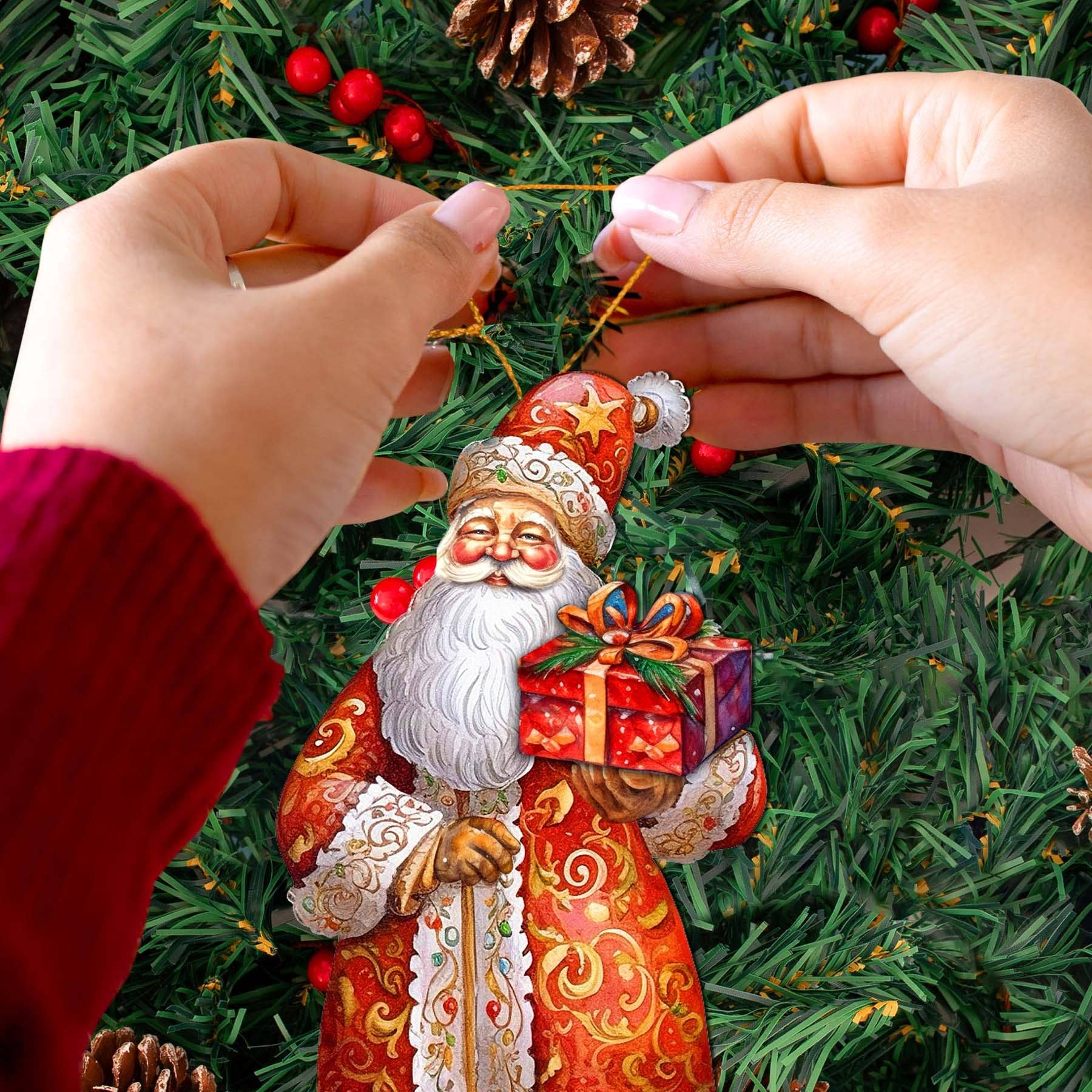Hands hang the St. Nick's Gift Wood Ornament by G Debrekht, a 5.5" x 3" Christmas Santa holding a wrapped present, on a decorated tree with pinecones and red berries.