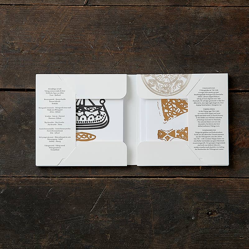 Open the Card Pack: Viking Finds Square Card Folder by Koustrup & Co - Denmark on a wooden surface to reveal intricate black and white Viking motifs with gold accents inside.