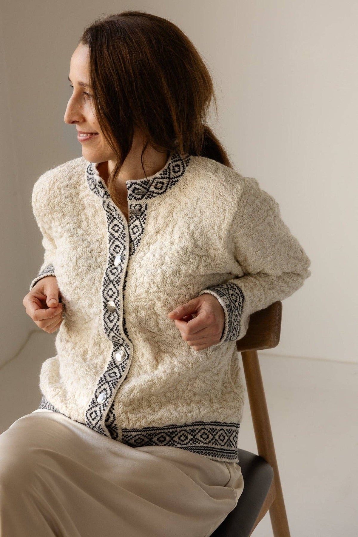 A woman sits on a chair, smiling to her left, wearing the cream-colored Bryggen Women's Norwegian Cardigan (White) with black geometric trim and a light skirt.