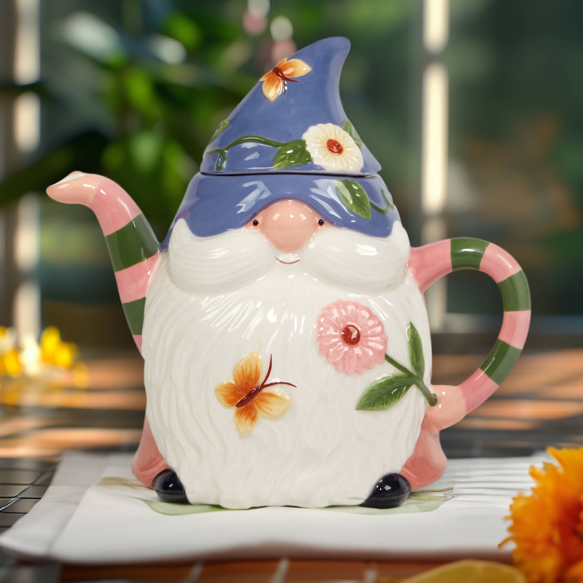 The Garden Gnomes 3-D Gnome Teapot (20oz), featuring a blue hat and floral accents, sits on a sunlit table.