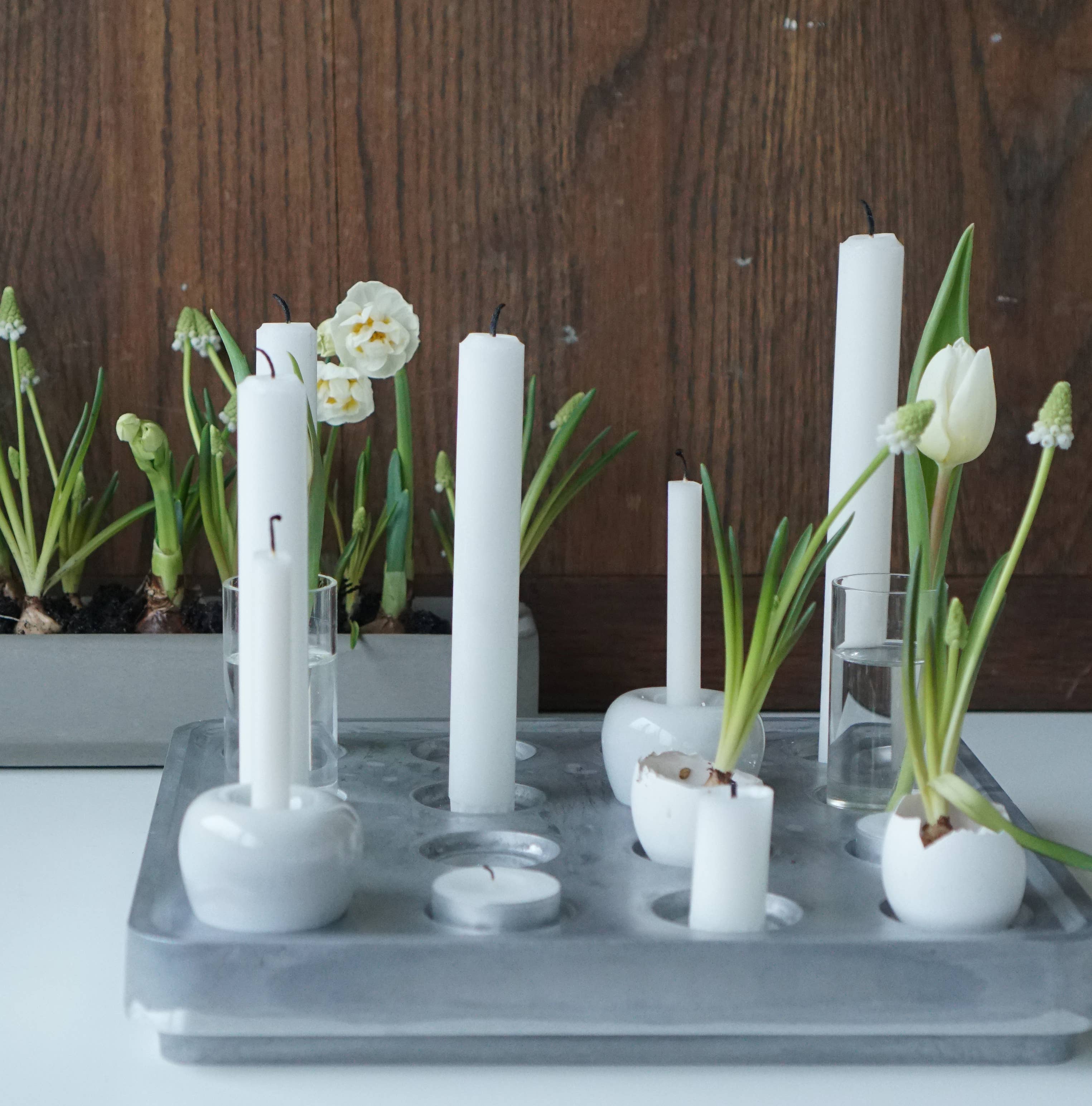 White taper candles and small white flowers in a minimalist stoneware holder from the Apple Candleholder 2-Pack Mini Apple, set against a brown wooden background with more flowers in a rectangular planter. The elegant design reflects Kim Vejsholts aesthetic.