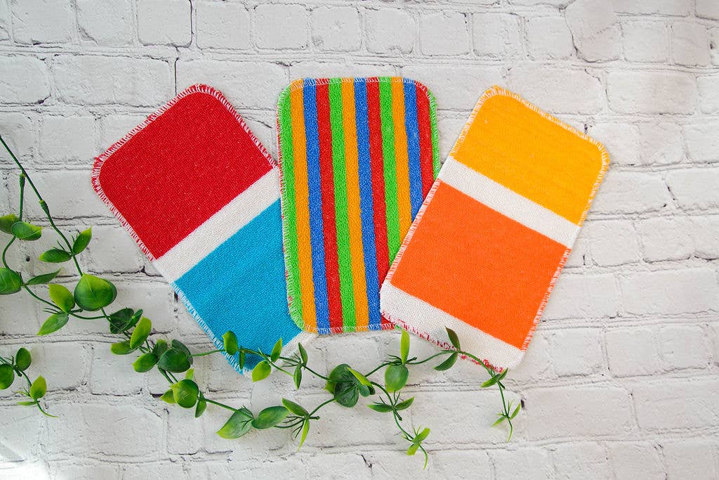 Three colorful rectangular fabric coasters with unique patterns are placed on a white brick surface next to a green vine and the Scrub: Scrub-Wow Extra Large Kitchen Cleaning Sponge.