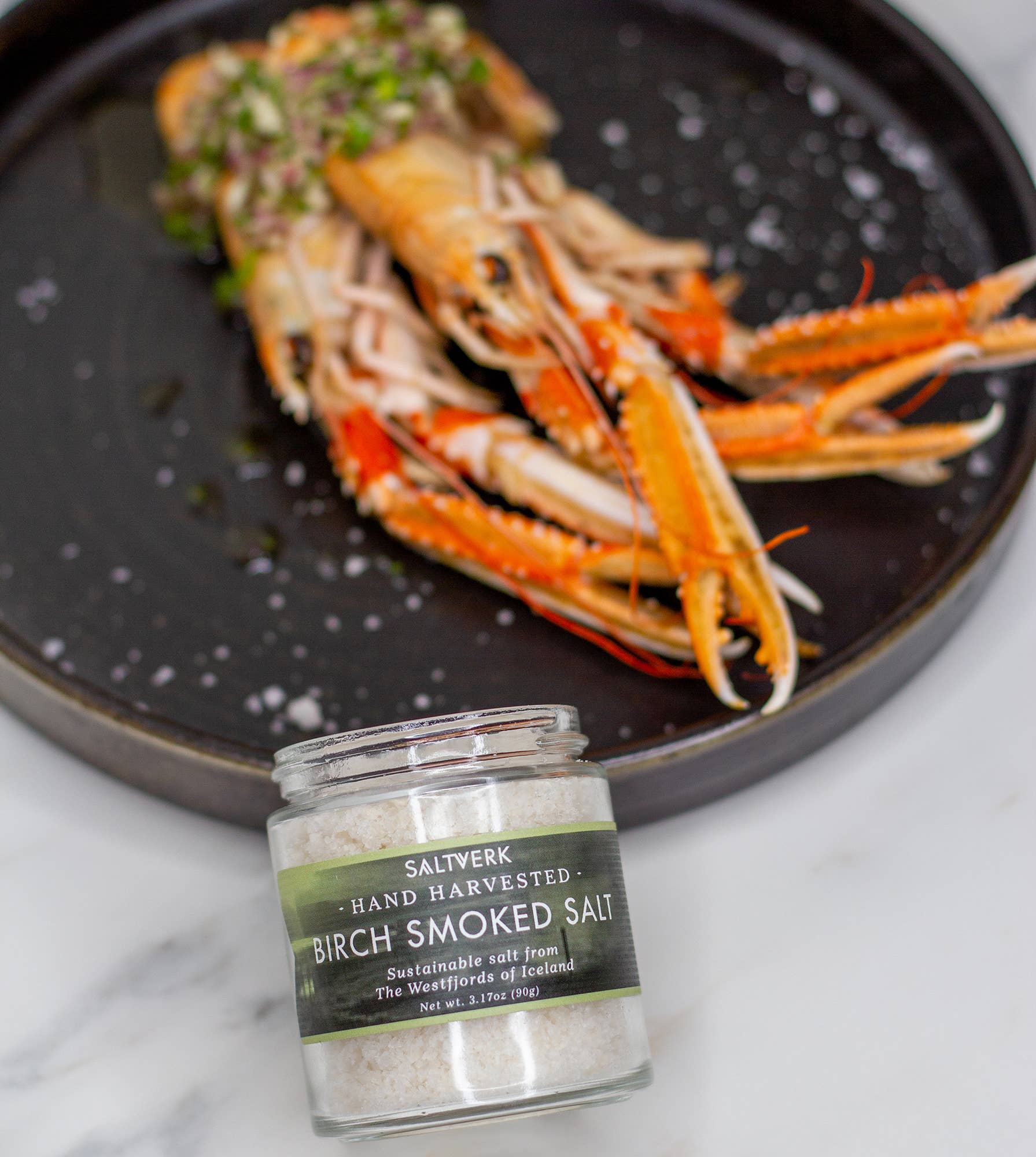 A plate of langoustines garnished with herbs sits behind the SALTVERK Gift Set, showing how these four Icelandic sea salts—Flaky, Lava, Birch Smoked, and Arctic Thyme—add rich flavor to seafood dishes.