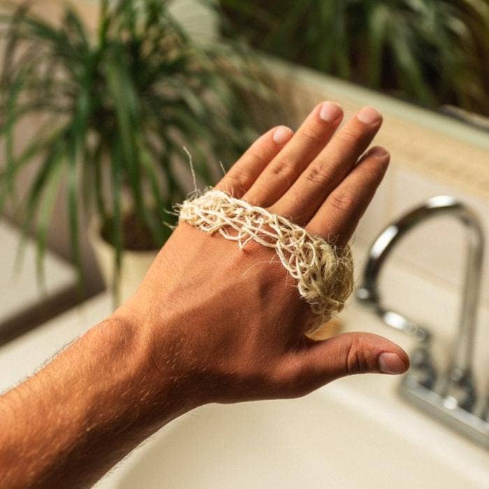 A hand holds the Scrub: Sisal Exfoliating Body Scrub by a bathroom sink, with a blurred plant in the background, showcasing this eco-friendly body scrubber perfect for zero-waste beauty routines.