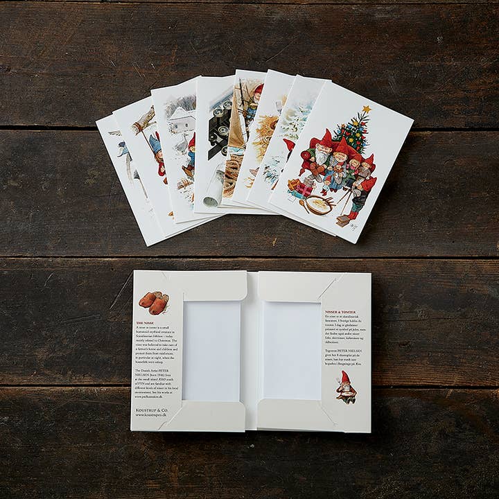 The "Nisser" Christmas Gnomes Card Pack by Koustrupco, featuring 8 unique images by Peter Nielsen with envelopes, is shown fanned out on a dark wood surface, with the open packaging visible below.