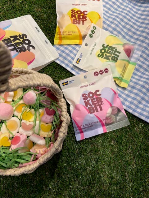 A wicker basket with colorful candy, including Sockerbit Sour Juicy Bottles Fruit Swedish Candy (2.8oz bags), sits on grass beside four open Sockerbit bags, with a blue and white checkered cloth in the corner.